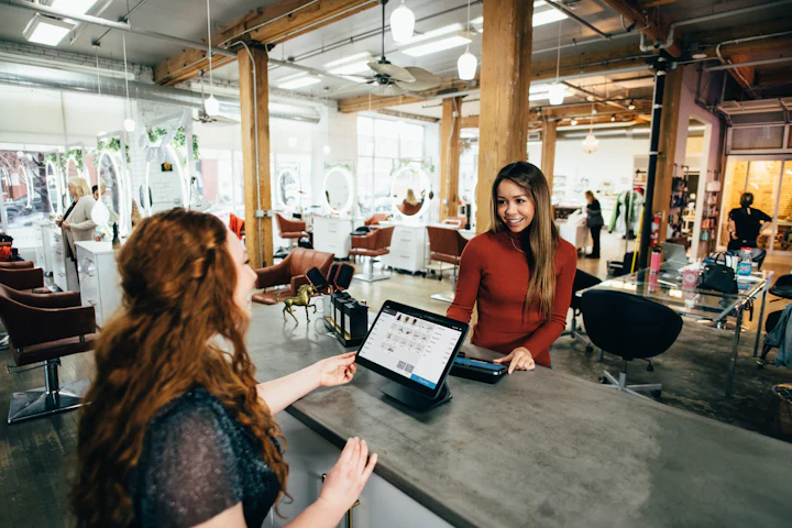 A salon owner smiling across the front counter as a customer pays at a tablet checkout.