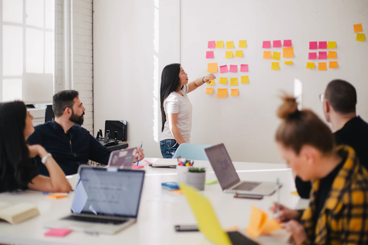 A small team planning around a meeting table while one person points to sticky notes on a whiteboard.